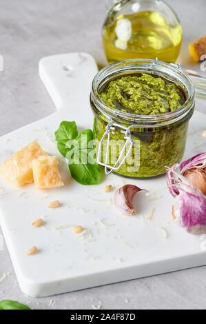 Il pesto o il pesto genovese in un vasetto di vetro con pinoli, parmigiano, basilico, olio e aglio su marmo bianco tagliere. Copia dello spazio. Foto Stock