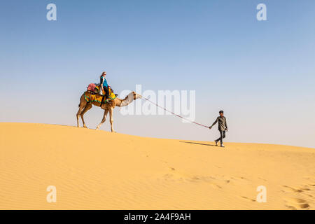 Un turista femminile su un cammello essendo guidato dal suo driver di cammello tra le dune di sabbia del deserto di Thar, Rajasthan, India. Foto Stock