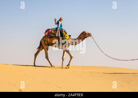 Un turista femminile su un cammello trek agitando per un'immagine in cima al suo cammello, il Deserto di Thar, Rajasthan, India. Foto Stock