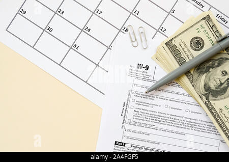 IRS Form W-9 richiesta per il contribuente un numero di identificazione e certificazione giace vuoto con penna e molte centinaia di fatture del dollaro nella pagina calendario. Imposta peri Foto Stock