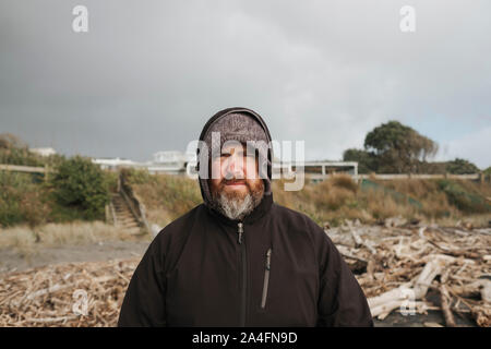 Uomo con barba permanente sulla spiaggia circondata da driftwood Foto Stock