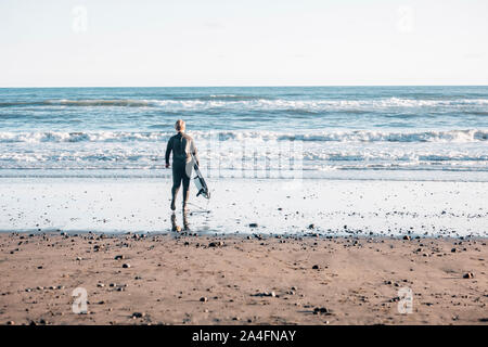 Tween boy in una muta in possesso di una tavola da surf a piedi in acqua Foto Stock