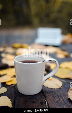 Su un vecchio tavolo di legno in autunno park è una tazza di tè e caffè, sparse in foglie di giallo e pigne. Vista dall'alto, in blur. Autunno Caldo buio Foto Stock