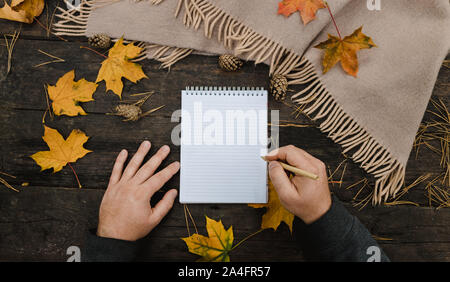 Le mani umane con una tazza di caffè e sciarpa al tavolo in legno con notebook e penna e foglie di autunno. Le mani umane con una tazza di caffè e sciarpa a Foto Stock