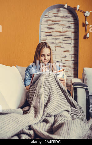 Ragazza seduta su un letto coperto con una coperta e studiando prima dell'esame, ella è la lettura di un libro e scrivere note con una matita Foto Stock