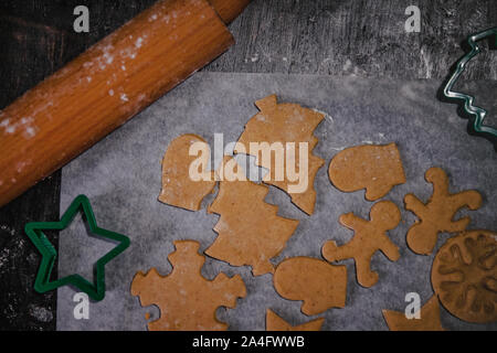 Tagliare gingerbread cookie nella forma di un albero di Natale, stella piccolo uomo, cuori da impasto crudo su pergamena carta da forno su uno sfondo scuro Foto Stock