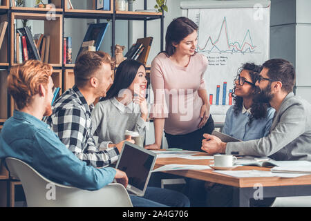 Startupers lavorando insieme a Office seduta strategia di sviluppo gioiosa guardando la donna incinta in piedi Foto Stock