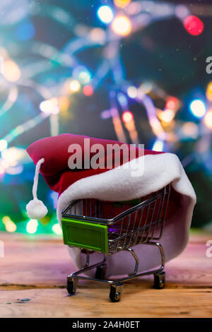 Shopping di Natale X-mas vendita concetto. Carrello spesa e Santa cap contro decorate con luci festose albero di Natale. Natale spendings. Inverno ho Foto Stock