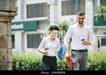 Felice sorridente giovane gente di affari a piedi in strada con tazze di caffè e le cartelle dopo la riunione di successo con investor Foto Stock