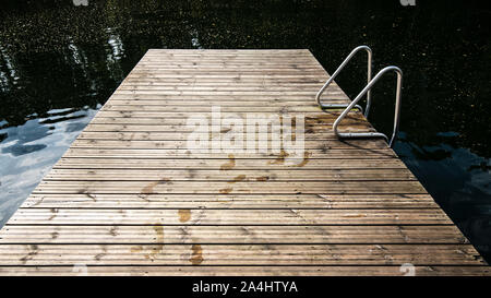 Un molo in legno con footprint di umido e una scala di metallo nel lago, contro lo sfondo di acqua calma, in un giorno di estate. Foto Stock