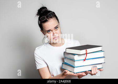 Giovane donna perplesso studente con gli occhiali sulla testa tiene una pila di libri nelle sue mani e si guarda attentamente alla fotocamera. Isolato su bianco backgrou Foto Stock