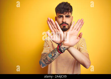 Giovane uomo con tatuaggio a strisce che indossa la t-shirt in piedi isolato su sfondo giallo rifiuto espressione bracci di attraversamento e di palme facendo sig negativa Foto Stock