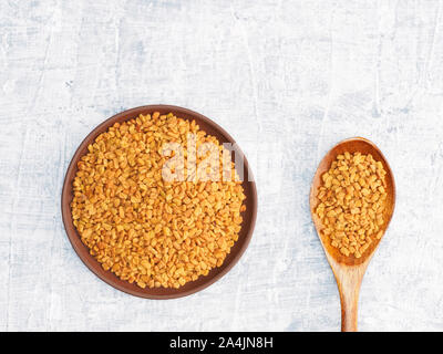 Spezie ed erbe contribuiscono a mantenere in buona salute e a migliorare l'appetito, vista dall'alto su sfondo di calcestruzzo. Fenugreek nella piastra di argilla e il cucchiaio di legno. Moderno Foto Stock