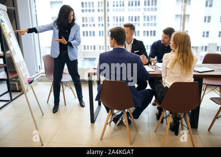 Clever brunette woman standing e presentando grafico sulla lavagna a fogli mobili durante il meeting aziendale, imprenditori e imprenditrici seduti al tavolo da conferenze a Foto Stock