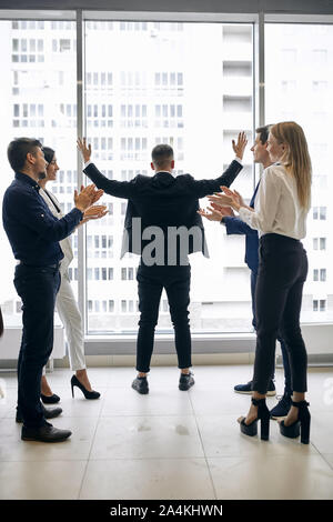 Successo imprenditore ambizioso di celebrare un trionfo con le braccia in alto, con il suo team ad applaudire il suo successo permanente, mentre in fron del panorama windo Foto Stock