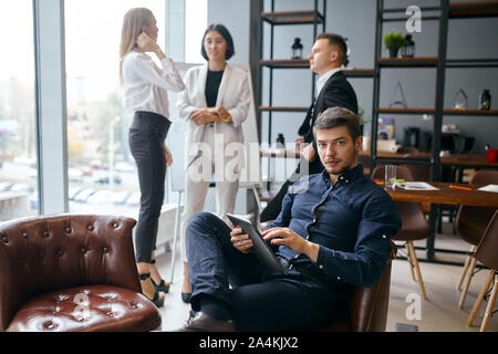 Attraente uomo in elegante maglietta blu avente un riposo sulla armschair mentre i suoi collaboratori avente una chat, conversazione aziendale, startup business Foto Stock