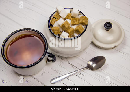 Il tè in un bianco smaltato, tazza di zucchero di canna e bianco a cubetti cubetti di zucchero in una ciotola di zucchero cucchiaino su una luce sullo sfondo di legno Foto Stock