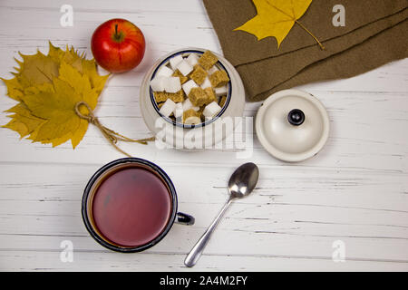 Il tè in una tazza bianca con zucchero bianco tagliato a cubetti e lo zucchero di canna, foglie di acero e mele su una tavola di legno sfondo luminoso. Foto Stock