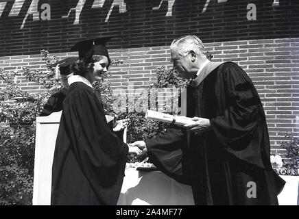 Anni '1960, storica, estiva e fuori alla cerimonia di laurea, una studentessa nel suo abito accademico e mortarboard che riceve il suo rotolo dal Cancelliere presso la University of Southern California (USC), Los Angeles, California, USA. Foto Stock