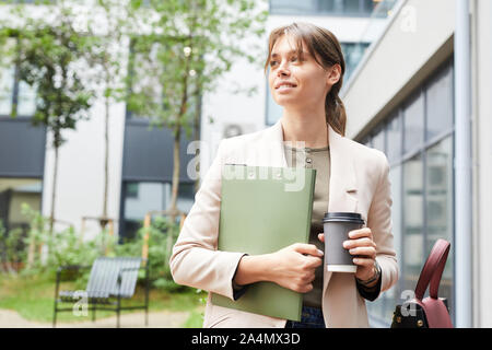 Giovane imprenditrice tenendo la cartella e tazza monouso di caffè nelle sue mani e passeggiate all'aperto nella città Foto Stock