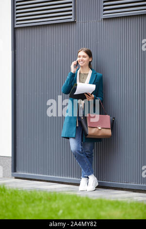 Giovane donna sorridente in elegante cappotto e con bella borsa appoggiata sulla parete e parlando al telefono cellulare nella città Foto Stock