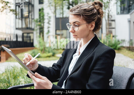 Malinconici business donna in cafe seduti sulla veranda hold digitale compressa e di bere succo di frutta. Bella donna che indossa gli occhiali sul lavoro digitale compressa in Foto Stock