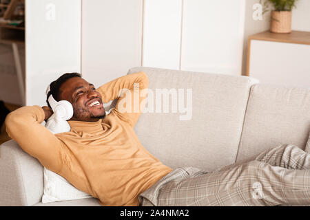 Afro-uomo in cuffie wireless sdraiato sul divano di casa Foto Stock