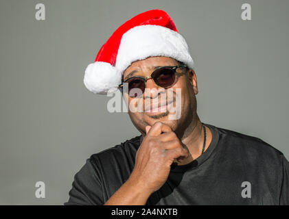 Sorridente African American uomo con la sua mano sul suo mento, indossando una di colore rosso e bianco Santa Claus hat contro uno sfondo a tinta unita Foto Stock