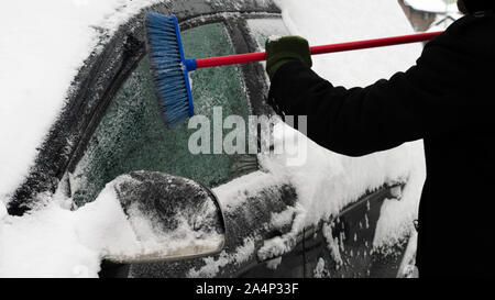 L'uomo cancella auto da neve in una fredda giornata invernale dopo la nevicata. Spazzola in mano mans. Tanta neve su auto. Foto Stock