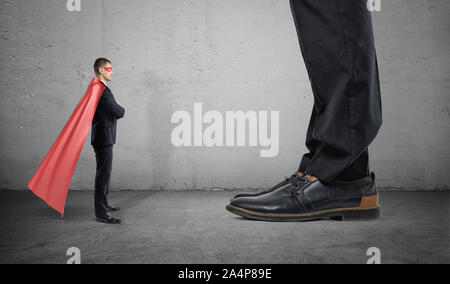 Un piccolo imprenditore in un supereroe cape sta di fronte l'uomo gigante con solo i suoi piedi visto. Foto Stock