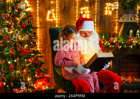 Santa Claus per aprire e leggere il libro magico con poco carino sorpreso ragazza in pigiama seduto sul ginocchio Foto Stock