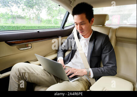 Giovane uomo asiatico utilizzando portatile sul sedile posteriore di una macchina Foto Stock