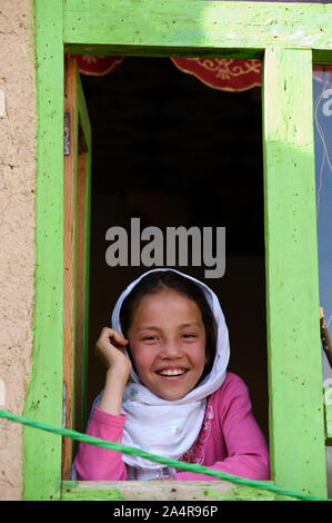 Un giovane afgano sorridente bambina guarda fuori dalla finestra di casa sua, nel villaggio di rag-e-Shad, nella periferia della città di Bamyan, centrale Provincia di Bamyan, Afghanistan. Il 11 maggio 2009. Foto Stock