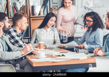 Startupers lavorando insieme a Office seduta guardando i documenti pensieroso mentre la donna incinta in piedi rivolta a informazioni importanti Foto Stock