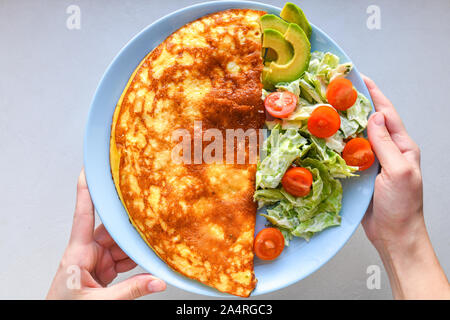 Uova fritte con verdure. le mani nel telaio. Frittata di vegetali su una piastra blu su un tavolo bianco, vista dall'alto Foto Stock