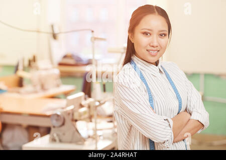 Ritratto di Asian giovane designer in piedi con le braccia incrociate e sorridente alla fotocamera in atelier Foto Stock