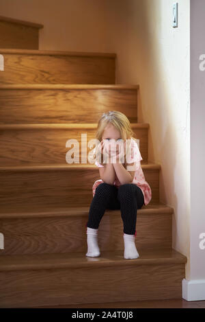 Bambina è seduta sulle scale per il secondo piano della casa, in attesa che i suoi genitori in solitudine. La luce del sole dalla finestra sta bruciando. Foto Stock