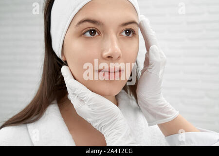 Mani di estetista in gomma bianca guanti medicali di toccare e ispezione volto del giovane, bella paziente nel cabinet di cosmetici. La donna in bianco bat Foto Stock