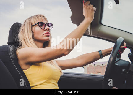 Una donna si ammira la sua bellezza nella riflessione. Allegro giovane donna alla guida di auto. Ragazza bionda guarda nello specchio e preens. donna abbassa la visiera parasole Foto Stock