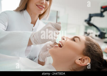 Vista laterale della bella donna sdraiata in poltrona del dentista. Un cliente contento denti di indurimento in odontoiatria. Professional dentista in guanti bianchi impedendo la carie, utilizzando strumenti di ripristino. Foto Stock