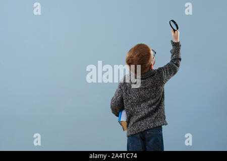 Curioso il ragazzo di zenzero in bicchieri guardando attraverso la lente di ingrandimento su blu Foto Stock