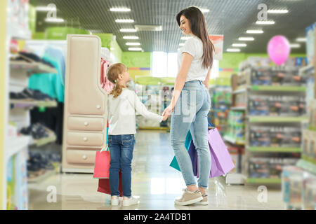 Vista dal retro del felice madre e figlia insieme per lo shopping nel centro commerciale. Giovani brunette mantenendo la mano della bambina, guardando il bambino e sorridente in negozio. Famiglia Acquisto di giocattoli per bambini. Concetto di acquisto. Foto Stock