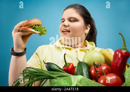Ritratto di ragazza in sovrappeso che finge di mangiare hamburger ma non la frutta e la verdura fresca che si tiene sul pacchetto verde. Felice femmina con mouse aperto è alla ricerca di un fast food oh la sua mano Foto Stock