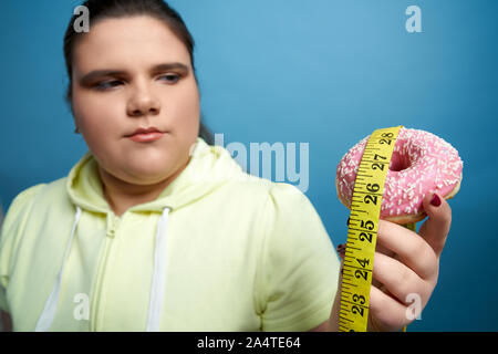 Close up plump brunette ragazze mano azienda dolce ciambella con smalto rosa e nastro di misurazione che pendono verso il basso. Bruciare i grassi e mangiare sano concetto Foto Stock