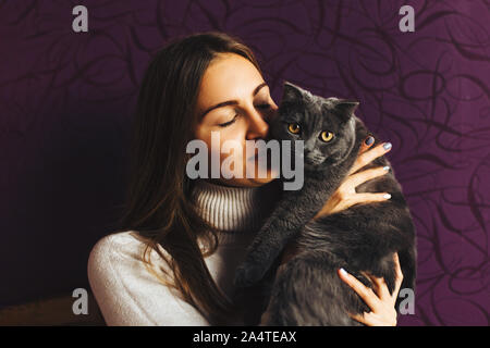 Bella ragazza giovane trattiene un soffice gatto grigio vicino al suo volto, home comfort e tranquillità e buon umore. Amore per gli animali Foto Stock