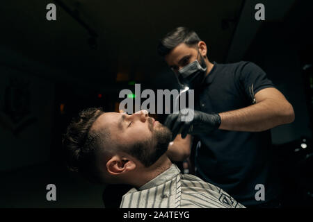 Vista laterale di uomo seduto in poltrona e coperto con abito a righe mentre il suo bel barbiere in guanti neri concentrati sulla barba per la rasatura con il rasoio. Concetto di servizio professionale al barbiere Foto Stock