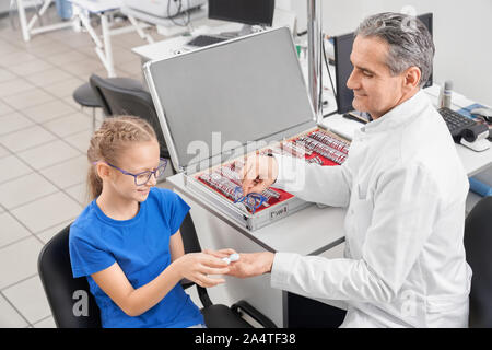 Vista da sopra di un anziano medico oculista maschio dando lente e bicchieri al piccolo paziente in clinica. Bella ragazza a parlare con il medico e la scelta di occhiali in negozio ottica. Concetto di oftalmologia. Foto Stock