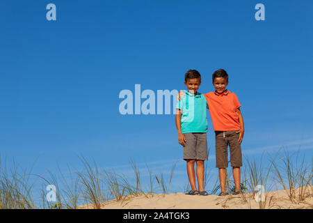 Due bello ragazzi piccoli stand sulla duna di sabbia su una spiaggia abbracciando insieme di indossare un abbigliamento informale oltre il cielo blu Foto Stock