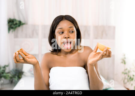 Funny divertente sorpreso americano africano giovane donna holding kiwi metà in mani su sfondo chiaro nella spa. Cura della pelle il concetto di bellezza. Foto Stock