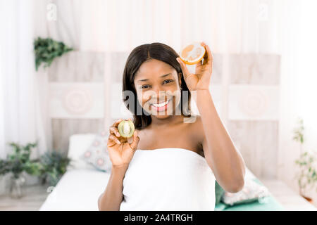 Funny divertente americano africano giovane donna holding kiwi e metà di colore arancione in mani su sfondo chiaro nella spa. Foto Stock